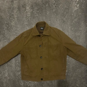 Brun jeansjacka från Zara - Säljer en brun jeansjacka från Zara med klassisk krage och knäppning framtill. Jackan har två stora bröstfickor och raka ärmar. Perfekt för dig som gillar en enkel och stilren look.