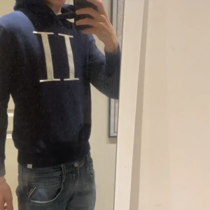 Mörkblå hoodie från Les deux - Snygg mörkblå hoodie från Les Deux med stor vit logga på bröstet. Klassisk huva och ribbade muddar. Perfekt för dig som gillar en stilren och sportig look. Passar till jeans eller joggers.