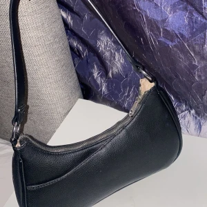 Svart axelväska från Don Donna - Snygg svart axelväska från Don Donna i stilren design. Väskan är gjord i strukturerat skinnimitation med silverfärgad dragkedja och ett ytterfack framtill. Perfekt storlek för det viktigaste och enkel att matcha till olika outfits.