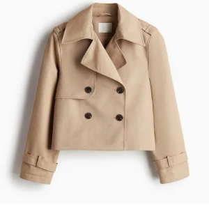Beige croppad trenchcoat med knappar - Snygg croppad trenchcoat i beige med dubbelknäppning framtill och bred krage. Jackan har långa ärmar med spänndetaljer vid handlederna. Storlek L men den är kort i modellen så det passar perfekt på mig som vanligtvis bär xs och s!