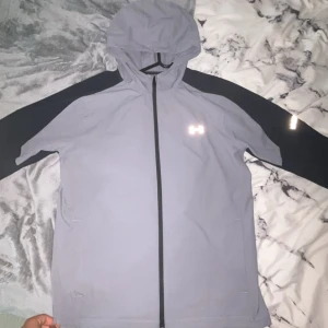 Under Armour vindjacka med huva - Snygg vindjacka från Under Armour i ljusgrått med svarta ärmar och hel dragkedja. Jackan har huva och reflekterande logga på bröstet, perfekt för blåsiga dagar. Tillverkad i lätt syntetmaterial och har en clean, sportig look. Finns även byxor till jackan