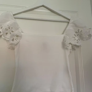Vit topp med volangärmar och spets - Superfin vit topp med breda volangärmar i spets och blommiga broderier. Toppen är ärmlös med axelband och har en rak, croppad passform. Perfekt för sommarens alla tillfällen och ger en romantisk vibe.