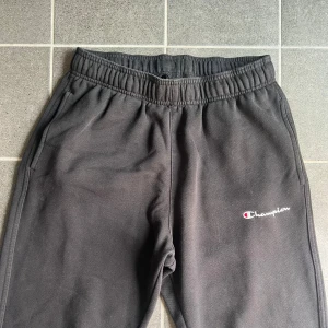 Svarta mjukisbyxor från Champion S - Svarta mjukisbyxor från Champion med broderad logga på benet. Byxorna har resår i midjan med snörning och är tillverkade i mjuk bomullsmix. Perfekta för chill eller träning, med normal passform och sidofickor.