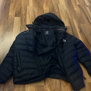 Svart pufferjacka Vinson Polo Club - Svart pufferjacka från Vinson Polo Club med huva och dragkedja. Jackan har broderade loggor på bröstet och ärmen samt coola detaljer med amerikansk flagga. Perfekt för kalla dagar och har en sportig vibe. Fodrad och quiltad för extra värme.