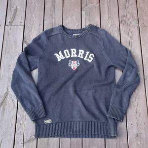 Mörkblå stickad tröja från Morris XS - Snygg mörkblå stickad tröja från Morris i storlek XS. Tröjan har vit broderad logga och emblem på bröstet samt ribbade muddar och nederkant. Perfekt för dig som gillar klassisk och sportig stil.