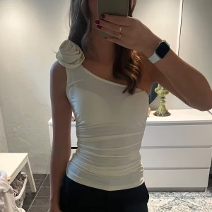 Benvit oneshoulder topp med blomdetalj - Säljer en benvit, figurnära oneshoulder topp med en stor tygblomma på axeln. Toppen har snygg rynkning längs sidorna och är tillverkad i ett stretchigt, glansigt material som ger en lyxig känsla. Perfekt för dig som vill sticka ut med en unik detalj på axeln.