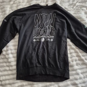 Svart Adidas sweatshirt med tryck - Svart sweatshirt från Adidas i bomull med stort grafiskt tryck på bröstet där det står 'NOW LOADING'. Tröjan har rund hals, långa ärmar och en relaxed passform. Perfekt för dig som gillar streetwear och gaming vibes.