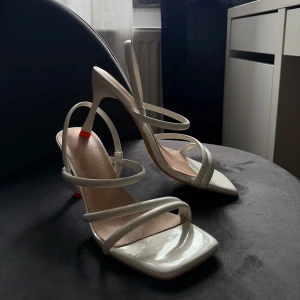 Vita sandaler med smala remmar och klack - Stilrena vita sandaler med smala remmar och fyrkantig tå. Skorna har hög klack och öppen tå. Snygg minimalistisk design med glansig finish och diskret detalj i rött under klacken.