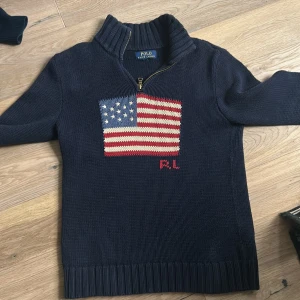 Mörkblå stickad tröja Polo Ralph Lauren - Mörkblå stickad tröja från Polo Ralph Lauren med halv dragkedja och hög krage. Framsidan har en stor amerikansk flagga i rött, vitt och blått samt RL-broderi. Perfekt för dig som gillar klassisk preppy stil och vill sticka ut.