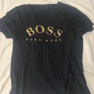 Svart Hugo Boss t-shirt med guldtryck - Snygg svart t-shirt från Hugo Boss med stort BOSS-tryck i guld på bröstet. Klassisk rund hals och korta ärmar. Perfekt för dig som gillar stilrena märkesplagg med lite extra attityd.