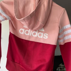 Adidas hoodie rosa och röd - Säljer en snygg Adidas hoodie i tvåfärgad design med ljusrosa upptill och mörkröd nedtill. Tröjan har huva, dragkedja och klassiska vita ränder på ärmarna. Perfekt för dig som gillar sportig stil och vill sticka ut lite extra.
