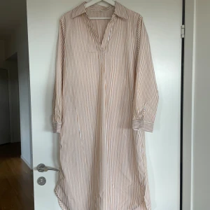 Randig långklänning med krage, storlek L - Snygg långklänning med orange/bruna och vita vertikala ränder. Klänningen har klassisk skjortkrage, v-ringad hals och långa ärmar med manschett. Materialet känns lätt och luftigt, perfekt för varmare dagar men även nu till hösten. Passformen är normal och modellen är rak.
