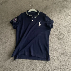 Marinblå polo t-shirt från Ralph Lauren - Snygg marinblå polo t-shirt från Ralph Lauren med vit logga på bröstet och Wimbledon-märke på ärmen. Klassisk krage med vita ränder och knappar framtill. Perfekt för dig som gillar sportig och stilren look.