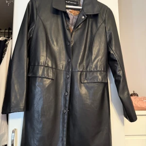 Svart skinnrock från Approlash - Snygg svart skinnrock från Approlash med coolt mönstrat innerfoder i orange och grått. Jackan har klassisk krage, knappar framtill och två stora fickor. Perfekt för dig som vill ha en stilren och edgy look.