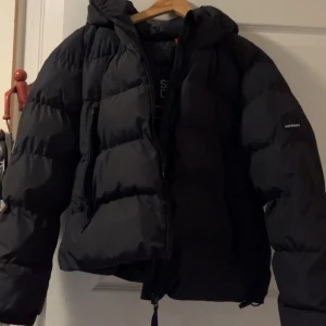 Svart pufferjacka från Superdry - Säljer en svart pufferjacka från Superdry med huva och dragkedja framtill. Jackan har en schysst oversized passform och är perfekt för kalla dagar. Fylld med syntetmaterial och har långa ärmar. Diskret Superdry-logga på ärmen. Använd max 3 gånger🩷🩷🩷