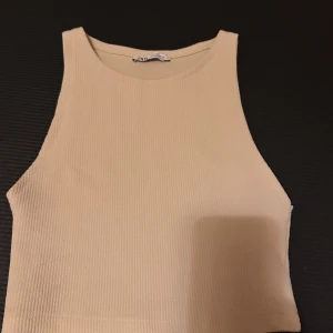 Beige ribbad linnetopp från Zara - Säljer en beige ribbad linnetopp från Zara. Toppen är croppad med hög halsringning och ärmlös design, perfekt för en clean och stilren look. Materialet är mjukt och stretchigt, vilket gör den superbekväm att bära. Passar snyggt till både jeans och kjol.