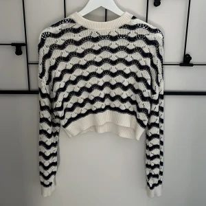 Vit och blå stickad tröja - Säljer en stickad tröja i vitt med blå vågiga ränder. Tröjan har lång ärm, ribbade muddar och en croppad passform. Perfekt för dig som gillar en chill och trendig look. Materialet är mjukt och luftigt, vilket gör den skön att bära under flera säsonger.