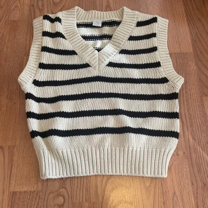 Randig stickad väst från Lindex - Snygg stickad väst från Lindex i storlek 134/140. Västen är beige med svarta horisontella ränder och har en bred ribbad kant nedtill och vid ärmöppningarna. V-ringad halsringning ger en klassisk look. Perfekt att slänga över en skjorta eller t-shirt.