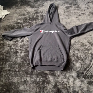 Grå hoodie från Champion, storlek L - Snygg grå hoodie från Champion i storlek L. Klassisk modell med broderad logga på bröstet, stor känguruficka och huva. Perfekt för chill dagar eller när du vill ha en enkel streetstyle-look. Mjuk och skön bomullsblandning inga snören 