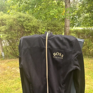 Svart Hugo Boss kofta  - Svart kofta från Hugo Boss med broderad logga i vitt på bröstet och ribbad krage med Hugo Boss-tryck. Kofta har hel dragkedja framtill och två fickor. Snygg, stilren och passar perfekt till streetwear eller chill dagar. Om ni vill se mer bilder adda mig på snap.