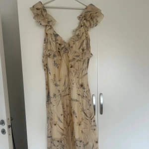 Blommig långklänning med volang från H&M -  Långklänning från H&M i ljus beige med blommigt mönster. Klänningen har volangdetaljer vid axlar och urringning, v-ringad fram och öppen rygg med knytning bak.