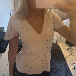 Prickig blus från Holly & Whyte XS - Söt ljusrosa blus med små svarta prickar från Holly & Whyte. Blusen har v-ringning, korta ärmar och markerad midja som ger en feminin siluett. Perfekt för dig som gillar en klassisk men ändå trendig look.