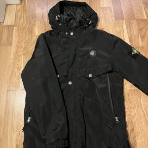 Svart parkajacka från Stone Island - Säljer en svart parkajacka från Stone Island med huva och flera fickor framtill. Jackan har dragkedja, tryckknappar och klassisk patch på ärmen. Materialet är glansigt och quiltat på insidan, perfekt för kyligare dagar. Snygg och funktionell design. som har många fickor.
