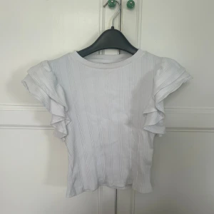 Vit ribbad topp med volangärmar Gina Tricot - Supersöt vit ribbad topp från Gina Tricot med volangärmar som ger en extra touch. Toppen har rund halsringning och är tillverkad i mjukt material, perfekt för en fräsch och stilren look. Passar perfekt till jeans eller kjol.