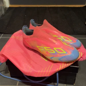 Adidas F50 rosa fotbollsskor - Säljer ett par Adidas F50 fotbollsskor i en riktigt fet rosa färg med gula och blå detaljer. Skorna har en strömlinjeformad design utan snörning och ovandel i syntetmaterial. Sulan är gul med blå inslag och dobbar för gräsplan. Perfekta för dig som vill sticka ut på planen!