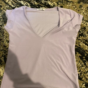 Ljuslila v-ringad topp från One Off - Säljer en ljuslila topp från One Off med v-ringning och korta ärmar. Toppen är enkel och stilren, perfekt att matcha med jeans eller kjol. Materialet känns mjukt och stretchigt, vilket gör den skön att bära hela dagen.