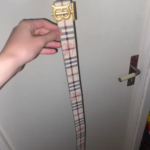 Burberry vändbart bälte med guldspänne - Säljer ett vändbart Burberry-bälte med klassiskt rutigt mönster på ena sidan i beige, svart. och svart med diskret rutmönster på andra sidan. Snyggt guldigt TB-spänne framtill. Bältet är gjort i skinn och passar perfekt till att lyfta din outfit.