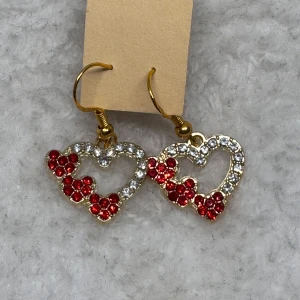 Glittriga hjärtformade örhängen - Snygga örhängen i hjärtform med glittrande vita och röda stenar. Basen är i guldfärgad metall och designen har flera små röda stenar som bildar ett mönster av hjärtan. Perfekt för dig som vill sticka ut med lite extra bling.