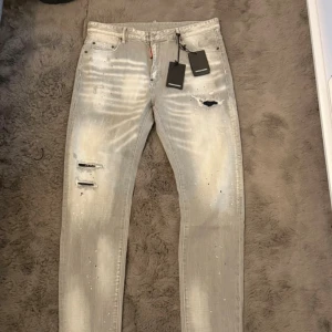 Grå distressed jeans från Dsquared2 - Säljer ett par grå jeans från Dsquared2 med coola slitningar, färgstänk och trasiga detaljer. Jeansen har en slim passform och klassisk femficksdesign. Materialet är jeans och färgen är ljusgrå med unika effekter. Perfekta för dig som gillar streetstyle. Dom är helt oanvända och fräscha.