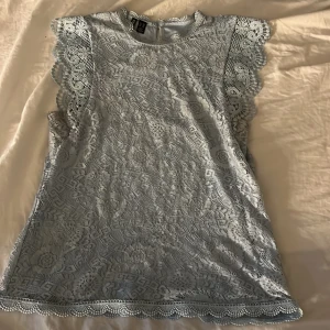 Ljusblå spetsblus från Pieces XS - Supersöt ljusblå blus från Pieces i storlek XS. Blusen har ett detaljerat spetsmönster med blommor och geometriska former, samt volangkant vid ärmar och nederkant. Den är ärmlös och har en liten knapp i nacken. Perfekt för dig som gillar romantisk och feminin stil.