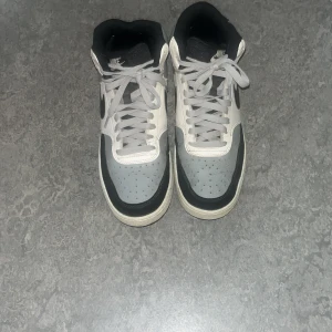 Nike grå och svarta sneakers high - Snygga Nike sneakers i high-modell med grå, svart och vit färgkombination. Skorna har klassisk snörning, rund tå och den ikoniska swoosh-loggan på sidan. Ovandelen är i syntet och textil, perfekt för dig som gillar streetstyle.