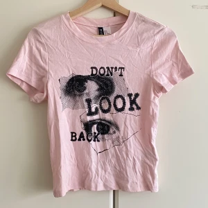 Rosa t-shirt med tryck från Divided - Snygg ljusrosa t-shirt från Divided med coolt svart tryck och texten 'DON'T LOOK BACK' samt grafiska ögon på framsidan. Modellen är klassisk med rund hals och korta ärmar, gjord i mjuk bomull. Perfekt för dig som gillar statement-plagg och streetstyle.
