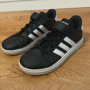 Svarta Adidas sneakers med vita ränder - Snygga svarta Adidas sneakers med klassiska vita ränder på sidorna och vit sula. Skorna har både snörning och kardborreband för extra smidig passform. Adidas-logga på häl och plös. Perfekta för en sportig och stilren look.