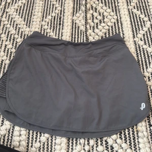 Grå sportkjol från Penn XS - Svart sportig kortkjol från Penn i storlek XS. Kjolen har inbyggda shorts under och är gjord i ett stretchigt syntetmaterial, perfekt för tennis eller träning. Enkel design med liten logga framtill och bekväm passform.