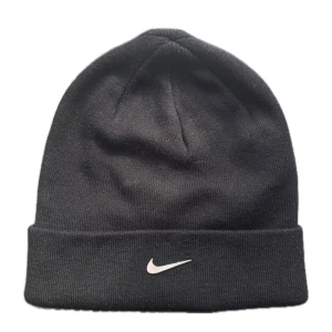 Svart mössa från Nike - Svart ribbstickad mössa från Nike med klassisk vikt kant och vit broderad swoosh-logga framtill. Perfekt för kyliga dagar och enkel att matcha med olika outfits. Tillverkad i mjukt och stretchigt material för skön passform.