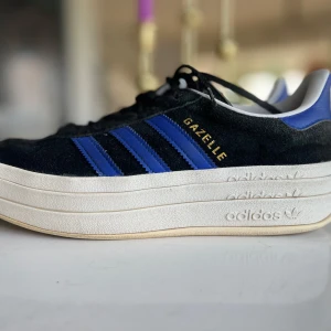Adidas Gazelle svart/blå sneakers 38,5 - Säljer ett par Adidas Gazelle sneakers i svart mocka med blå detaljer och klassiska tre ränder på sidan. Skorna har en chunky vit plattformssula med Adidas-logga, snörning och vit insida. Perfekta för dig som gillar retrostil och vill sticka ut.