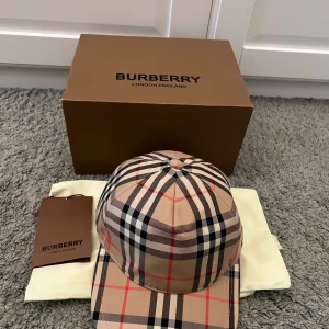 Burberry rutig keps - Säljer en klassisk rutig keps från Burberry i beige, svart, röd och vit. Kepsen har justerbar passform och är tillverkad i polyester. Perfekt accessoar för en stilren look.