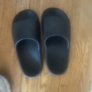 Svarta sandaler i syntet - Chunky svarta sandaler med bred rem och platt sula. Tillverkade i syntetmaterial och har en minimalistisk design som passar perfekt till avslappnade outfits.