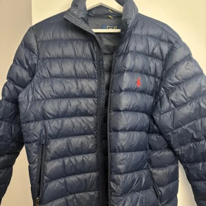 Blå dunjacka från Polo Ralph Lauren - Snygg blå dunjacka från Polo Ralph Lauren med klassisk quiltad design och dragkedja framtill. Jackan har två fickor med dragkedja och den ikoniska röda loggan broderad på bröstet. Perfekt för kyliga dagar och riktigt stilren look.