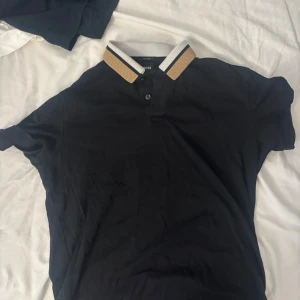 Svart pikétröja från Hugo Boss - Svart pikétröja från Hugo Boss med klassisk krage i beige, vitt och svart. Tröjan har korta ärmar och två knappar vid halsen. Materialet känns mjukt och är troligtvis bomull, perfekt för en clean och stilren look.