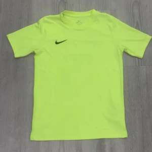Neongul Nike Dri-Fit fotbollst-shirt - Neongul t-shirt från Nike med Dri-Fit-teknologi, perfekt för fotboll. Kortärmad och med svart Nike-logga på bröstet samt tryck på ryggen med Stadium Sports Camp och FOTBOLL. Skön passform och lätt material som andas.