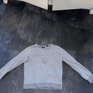 Grå sweatshirt från GANT - Snygg ljusgrå sweatshirt från GANT med broderad logga och sköld framtill. Tröjan har rund halsringning, långa ärmar och ribbade muddar. Perfekt för en chill och stilren look, passar till jeans eller mjukisbyxor.