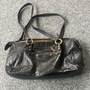  Vintage❤️ - Stilren svart axelväska i krokopräglat skinn med snygga guldfärgade metalldetaljer. Väskan har flera fack, dragkedja upptill och en rymlig insida med beige foder. Perfekt för dig som vill ha en edgy och klassisk look. Lite fläckar inne se sista bild❤️