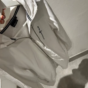 Vit hoodie från Sail Racing med dragkedja - Vit hoodie från Sail Racing med hel dragkedja och extra bröstficka med dragkedja. Broderad logga på bröstet och svartvit randig detalj på insidan av kragen. Mjuk insida i frottéliknande material och huva för extra chill vibe.