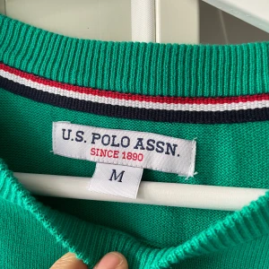Grön stickad tröja från U.S. Polo  - Snygg grön stickad tröja från U.S. Polo Assn. Tröjan har rund halsringning med ribbade detaljer och långa ärmar. Perfekt för dig som gillar klassisk och stilren look.