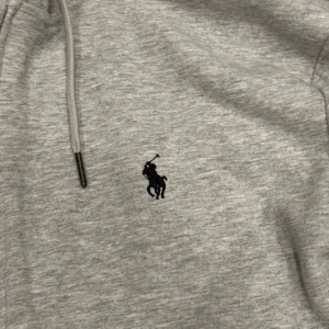 Grå hoodie från Polo Ralph Lauren - Snygg ljusgrå hoodie från Polo Ralph Lauren med dragkedja och huva. Tröjan har långa ärmar, snörning i huvan och klassisk broderad logga på bröstet. Tillverkad i mjuk bomull, perfekt för chill dagar eller när du vill ha en clean look.